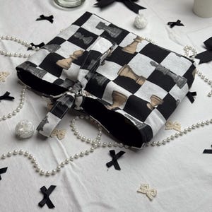 Può includere: Una borsa a scacchi in bianco e nero con un motivo a pezzi degli scacchi. La borsa è legata con un fiocco di tessuto abbinato. Fiocchi neri decorativi, perle e piccoli fiocchi bianchi sono sparsi intorno alla borsa su una superficie bianca.