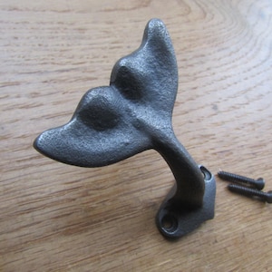 May include: A dark gray metal wall hook shaped like a whale tail with a textured, matte finish. The hook features a curved neck and a flat mounting base with two screw holes. Two small black metal screws are positioned next to the hook.