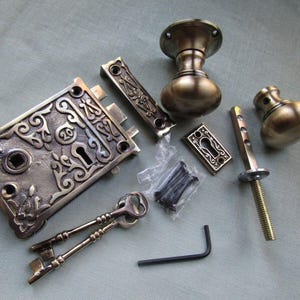 Peut inclure: Un ensemble de quincaillerie de porte en laiton comprenant une plaque de serrure à mortaise rectangulaire avec des volutes ornées et le numéro 20, deux poignées rondes, une rosace de serrure, deux clés, une tige filetée, une clé Allen et des vis.
