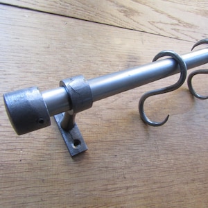 May include: A dark grey metal rod featuring a cylindrical end cap and a wall-mounted bracket. Two S-shaped metal hooks are suspended from the rod. The metal has a textured, matte finish with visible signs of craftsmanship on the bracket and hooks.