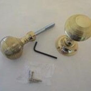 Op de afbeelding: Twee deurknoppen van gepolijst messing met een bolvormig, geribbeld ontwerp. Eén knop is voorzien van een metalen spindel, terwijl de andere op een ronde voetplaat is gemonteerd. Een zwarte inbussleutel en een zakje met schroeven zijn inbegrepen.