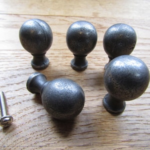 May include: A set of five spherical cabinet knobs with a dark, textured, metallic finish and a single silver-toned metal mounting screw. Each knob features a rounded head attached to a tapered base, showcasing a consistent, weathered appearance.