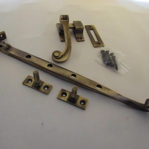 May include: A set of antique brass window hardware including a long casement stay with multiple adjustment holes, a curved sash fastener, two mounting pins, a rectangular strike plate, and a small plastic bag containing several black screws.