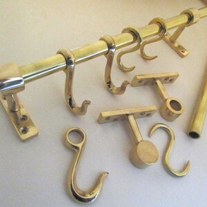 May include: A polished brass rail system featuring a horizontal rod with square end caps, multiple S-shaped hooks hanging from the rod, and two wall-mounted brackets with circular supports, accompanied by an additional separate brass rod.