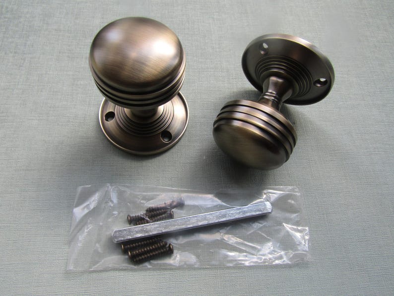 K&ouml;nnte beinhalten: Zwei runde T&uuml;rkn&ouml;pfe mit geb&uuml;rstetem Bronze-Finish, die ein geripptes Design am Hals und an der Basis aufweisen. Das Set enth&auml;lt einen durchsichtigen Plastikbeutel mit einem Metallstift und mehreren kleinen Befestigungsschrauben zur Montage.