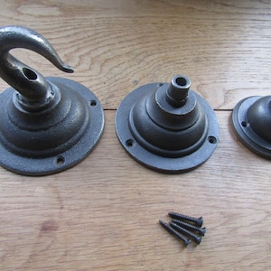 Könnte beinhalten: Drei dunkelgraue Deckenhaken und Halterungen aus Metall in verschiedenen Größen, jeweils mit einer kreisförmigen Basis mit vorgebohrten Schraublöchern. Das Set enthält einen großen Hakenaufsatz und zwei kleinere kuppelförmige Halterungen sowie vier schwarze Schrauben.
