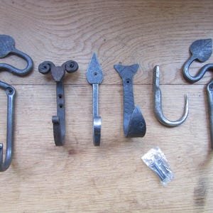 May include: A collection of six dark gray metal wall hooks with varied designs, including two with leaf-shaped tops, one with a ram head, one with a pointed spearhead, one with a fish tail, and one simple J-hook, accompanied by a small bag of screws.