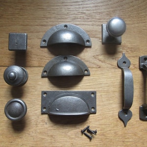 Puede incluir: Una colección de herrajes de metal gris oscuro que incluye tres tiradores de media luna, dos manijas verticales con extremos decorativos, tres pomos redondos, un pomo cuadrado y dos tornillos negros pequeños sobre una superficie de madera.