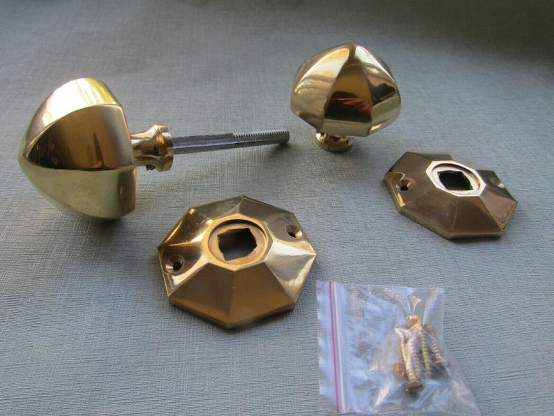 Puede incluir: Dos pomos de puerta de lat&oacute;n pulido con formas geom&eacute;tricas facetadas, acompa&ntilde;ados de dos placas octogonales a juego y una peque&ntilde;a bolsa de pl&aacute;stico que contiene varios tornillos de montaje de metal. Acabado en color dorado.