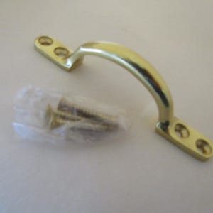 Puede incluir: Un tirador de mueble de latón pulido con un diseño de arco curvado y dos placas de montaje planas, cada una con dos orificios para tornillos preperforados. Una pequeña bolsa de plástico transparente con varios tornillos de latón a juego se sitúa al lado.