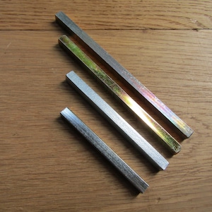 May include: Four metal door handle spindles of varying lengths arranged diagonally. The spindles have a square cross-section and feature different finishes, including a dull silver tone and a reflective, iridescent gold-toned metallic surface.