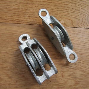 May include: Two silver-colored metal pulleys. One pulley features a single wheel housed within a frame with an eyelet at each end. The second pulley features a double wheel configuration within a similar metal frame with an eyelet at one end.