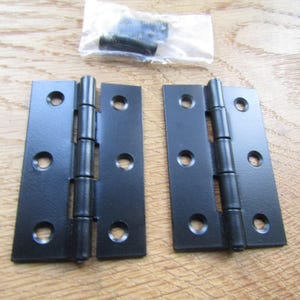 May include: Two rectangular matte black metal butt hinges, each featuring three circular screw holes on both sides of the central barrel. A small clear plastic bag containing black metal screws is positioned above the two hinges.