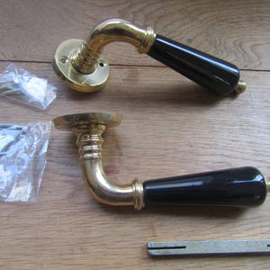 May include: Two door handles featuring polished brass bases and curved necks with glossy black grips. The set includes two small plastic bags containing mounting screws and an Allen key, along with a single metal spindle bar for installation.