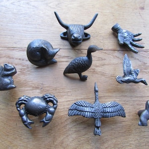 May include: A collection of nine dark gray metal cabinet knobs featuring various shapes including a bull head, a human hand, a curled cat, a standing duck, a squirrel, a crab, a flying bird, a bird with wings spread, and a small dog figure.