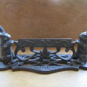 May include: A dark grey cast iron boot scraper featuring two upright rabbits facing each other, holding a central horizontal bar with decorative scrollwork. The base is rectangular with rounded ends and a lattice pattern, providing a stable foundation.