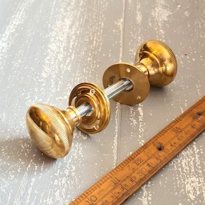 Könnte beinhalten: Ein Türknauf-Set aus poliertem Messing mit zwei abgerundeten Knäufen mit gerippten Kanten, die durch einen zentralen Metallstift und zwei kreisförmige Montageplatten verbunden sind. Ein Holzlineal liegt daneben und zeigt Maße in Zentimetern.