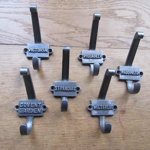 Könnte beinhalten: Ein Set aus fünf dunkelgrauen Wandhaken aus Metall, jeder mit einer rechteckigen Platte und einem erhabenen Namen. Die Namen sind Victoria, Piccadilly, Paddington, Covent Garden und St Pancras. Jeder Haken hat zwei Schraublöcher zur Montage.