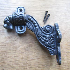 Puede incluir: Un soporte de pared de metal gris oscuro con un diseño curvo y ornamentado que presenta volutas y una cabeza de animal esculpida en la parte superior. El soporte incluye dos placas de montaje con orificios y dos tornillos metálicos negros.