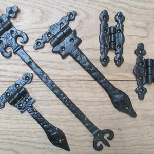 May include: A collection of five black metal hinges with a textured finish. The set includes two long strap hinges with decorative ends, two smaller hinges with ornate cutouts, and one rectangular hinge with rounded edges and multiple screw holes.