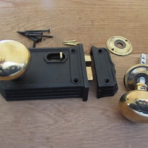 May include: A black rim lock set featuring two polished brass round knobs, a rectangular black lock body, a matching black strike plate, a circular brass escutcheon, and a collection of small black and brass mounting screws for installation.