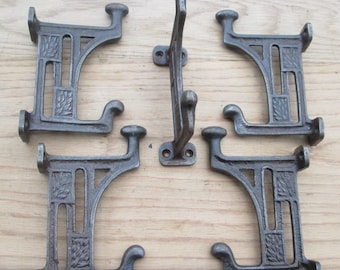 Könnte beinhalten: Ein Set aus fünf Wandhaken aus dunkelgrauem Gusseisen. Vier Haken sind in einer rechteckigen Formation angeordnet, jeder mit einem durchbrochenen Rahmen mit geprägten Blattmustern und zwei Zinken. Ein Haken ist mittig vertikal platziert.
