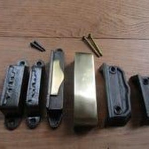 May include: A set of metal window sash locks and hardware components including two dark cast iron latch bodies, a brass-coloured latch plate, a polished brass sash lift, and two small black screws alongside two longer brass-coloured mounting screws.