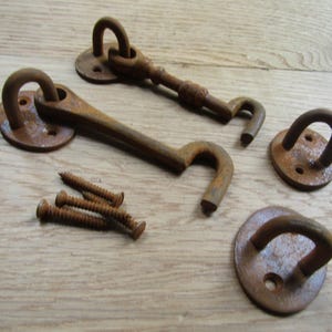 May include: Two rusty metal cabin hooks with matching circular mounting plates and three matching screws. Each hook features a curved arm attached to a round base with two screw holes, displaying a weathered, oxidized brown finish throughout.