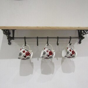 May include: A wall-mounted shelf featuring a light-toned wooden board supported by two dark metal brackets. A horizontal metal rod is attached beneath the shelf, holding five curved hooks. Three white ceramic mugs with red and grey floral patterns hang from the hooks.