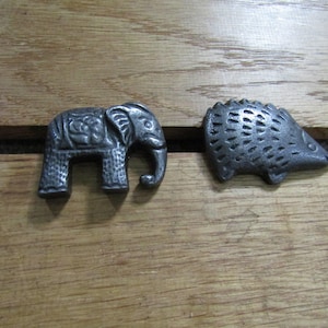 May include: Two small, dark grey metal figurines shaped like animals. One figurine is an elephant with textured details on its body and ear. The other figurine is a hedgehog with a spiky, textured back and a pointed snout, both resting side by side.