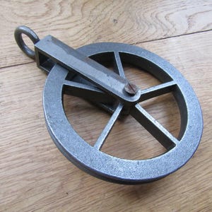 May include: A grey metal pulley featuring a circular wheel with six spokes held within a rectangular frame. A single metal eyelet is attached to the top of the frame, and a central screw secures the wheel to the structure, showing a textured finish.