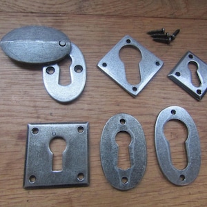 Könnte beinhalten: Eine Sammlung silberfarbener Schlüsselloch-Rosetten aus Metall in verschiedenen Formen wie oval, quadratisch und rautenförmig, mit vorgebohrten Schraublöchern. Das Set enthält eine passende ovale Abdeckplatte und kleine schwarze Metallschrauben.