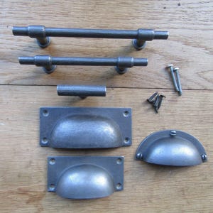 May include: A collection of dark grey metal cabinet hardware including two long bar pulls, one T-shaped knob, two rectangular cup pulls, one semi-circular cup pull, and a small assortment of matching metal screws for installation purposes.