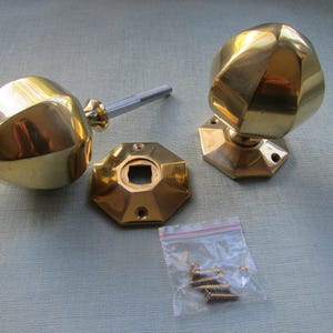 K&ouml;nnte beinhalten: Zwei polierte T&uuml;rkn&ouml;pfe aus Messing mit facettierten, geometrischen Formen, jeweils an einer passenden achteckigen Grundplatte befestigt. Ein Knauf enth&auml;lt einen Metallstift. Ein kleiner durchsichtiger Plastikbeutel enth&auml;lt mehrere Schrauben.