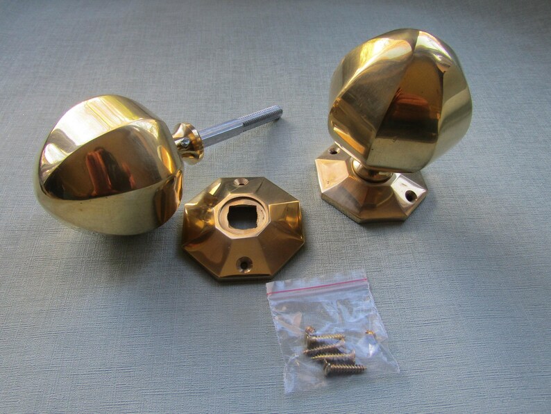 K&ouml;nnte beinhalten: Zwei polierte T&uuml;rkn&ouml;pfe aus Messing mit facettierten, geometrischen Formen, jeweils an einer passenden achteckigen Grundplatte befestigt. Ein Knauf enth&auml;lt einen Metallstift. Ein kleiner durchsichtiger Plastikbeutel enth&auml;lt mehrere Schrauben.