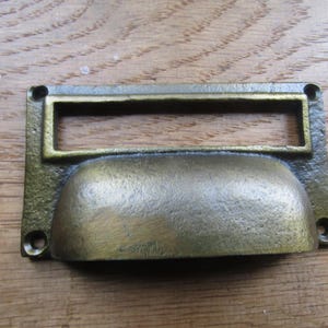 May include: A rectangular brass drawer pull with a cup-shaped handle and an integrated label holder slot. The hardware features a weathered, dark metallic finish and includes four small circular mounting holes at each corner for attachment.