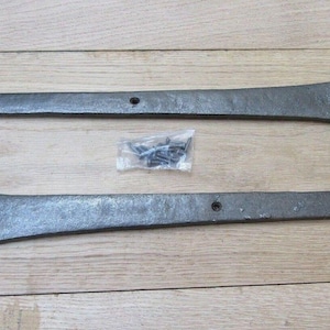 Op de afbeelding: Twee lange, donkergrijze metalen hengen met een gehamerde textuur en een puntig, speervormig uiteinde. Elke heng heeft een rechthoekige montageplaat met voorgeboorde gaten en een centrale scharnierpen. Een zakje met schroeven is inbegrepen.