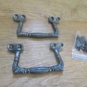 Könnte beinhalten: Zwei passende silberfarbene Metall-Schubladengriffe mit texturiertem, verziertem Design und zwei Befestigungslöchern an jedem Ende. Ein kleiner durchsichtiger Plastikbeutel mit mehreren dunklen Metallschrauben liegt rechts neben den Griffen.