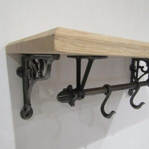 May include: A wall-mounted shelf featuring a light-coloured wooden board supported by two ornate, dark metal brackets. A horizontal metal rod is attached to the brackets, equipped with two movable S-shaped hooks for hanging items.