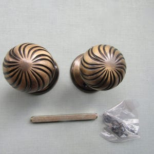 May include: Two round brass door knobs featuring a swirling, radial ribbed pattern with dark recessed grooves. A single rectangular metal spindle bar and a small clear plastic bag containing several dark metal screws are included in the set.