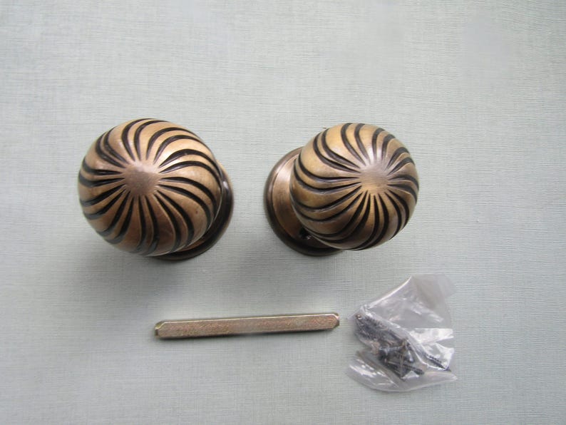May include: Two round brass door knobs featuring a swirling, radial ribbed pattern with dark recessed grooves. A single rectangular metal spindle bar and a small clear plastic bag containing several dark metal screws are included in the set.