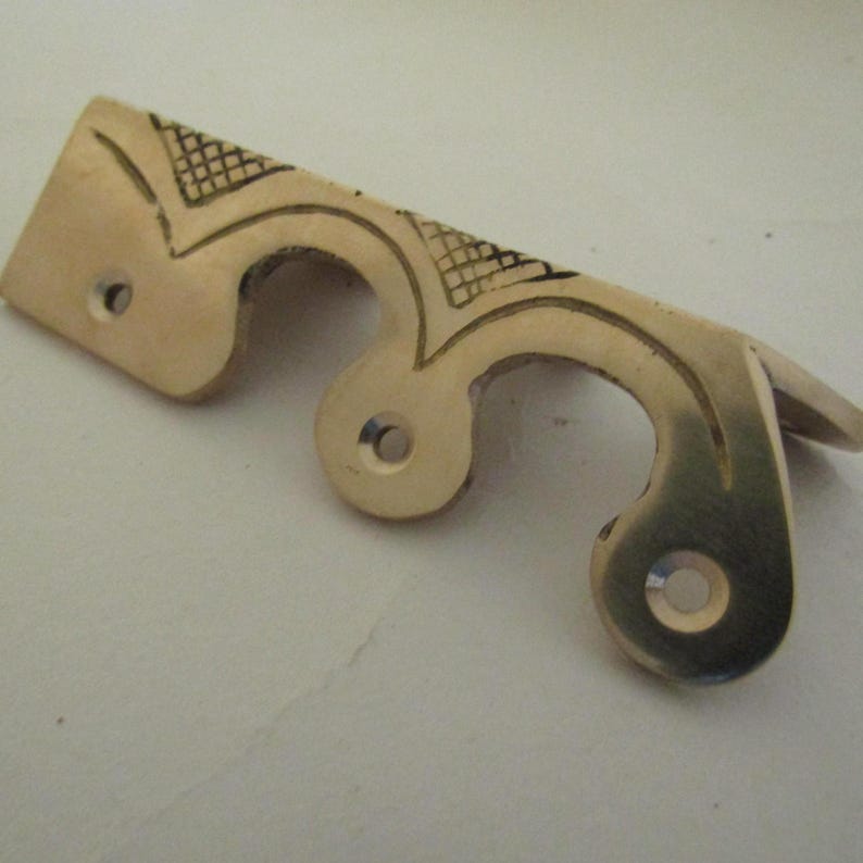 May include: A polished brass furniture pull featuring a curved, scalloped edge design with three circular mounting holes. The metal surface is decorated with engraved cross-hatched patterns and smooth, flowing lines along the top edge.