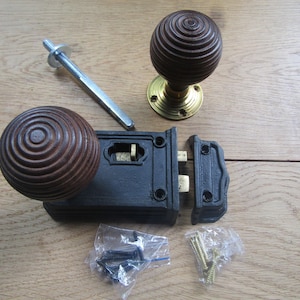 May include: A rim lock set featuring two dark brown spherical wooden knobs with concentric ring patterns, a black metal lock body, a brass mounting plate, a metal spindle, and two small plastic bags containing black and gold mounting screws.