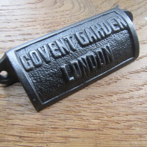 May include: A dark grey metal cup pull handle with a textured surface and raised lettering that reads GOVENT GARDEN LONDON. The hardware features two screw holes on the side flanges for mounting to a drawer or cabinet front surface.
