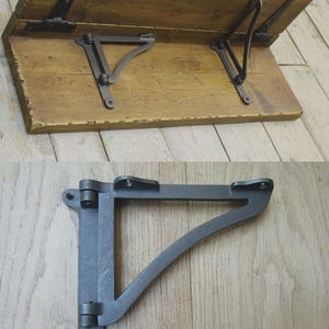 May include: A wooden shelf with a natural finish and distressed edges, featuring two black metal folding brackets attached to the underside. Below, a single black metal triangular folding shelf bracket with a hinge mechanism and screw holes.