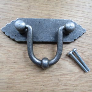 Könnte beinhalten: Ein dunkelgrauer Schubladengriff aus Metall mit einer rechteckigen Rückplatte mit gewellten Kanten und einem U-förmigen Klappgriff. Das Zubehör umfasst zwei silberfarbene Metallschrauben mit Gewindeschäften zur Montage an Möbeln.