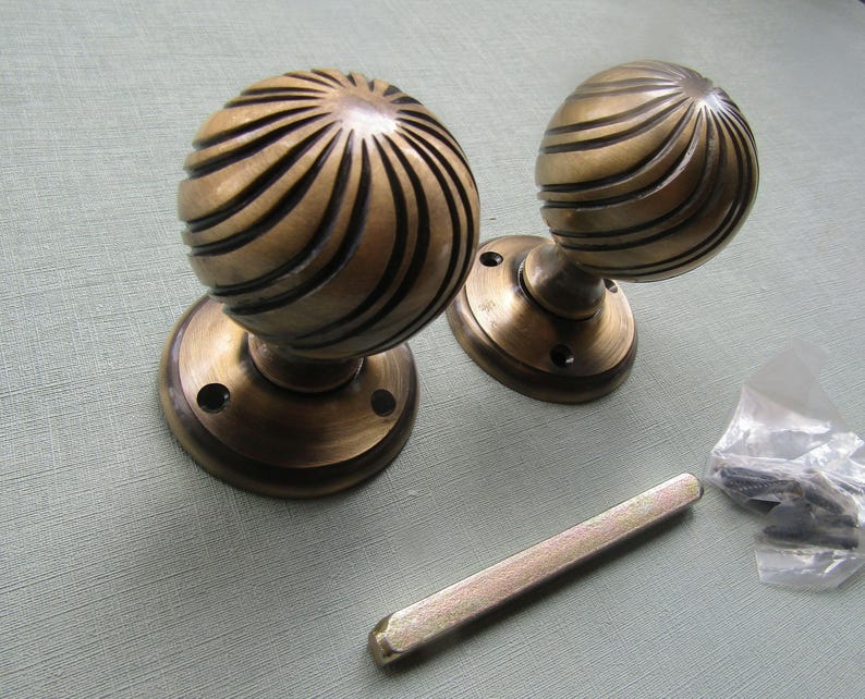 May include: Two round brass door knobs featuring a spiraling ribbed texture and a dark antique finish. Each knob is attached to a circular mounting plate with screw holes. A single metal spindle bar and a small plastic bag of screws are included.