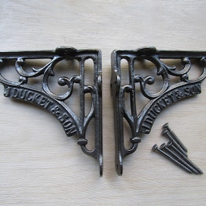 May include: Two dark gray cast iron shelf brackets with an ornate scrollwork design and the embossed text J DUCKET & SON. The set includes four matching metal screws positioned next to the right bracket, featuring mounting holes for installation.