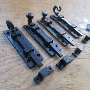 May include: Four black metal door bolts with textured surfaces, each featuring a sliding latch mechanism and a knob handle. The set includes four matching mounting brackets and a small plastic bag containing black screws for installation purposes.