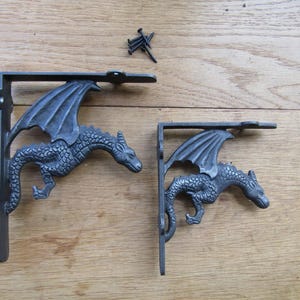 Puede incluir: Dos soportes de estante de metal gris oscuro con un diseño de dragón que presenta escamas y alas texturizadas. Los soportes tienen marcos en forma de L con orificios de montaje y vienen acompañados de varios tornillos negros para su instalación.